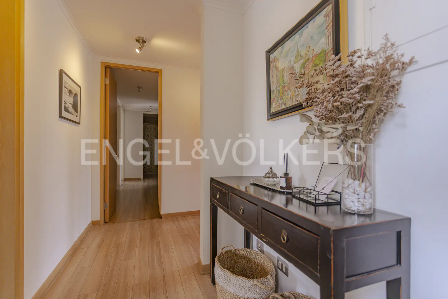 Hallway with light wood floors, white walls, and a dark wood console table with a vase of dried flowers. A framed painting hangs above the table.