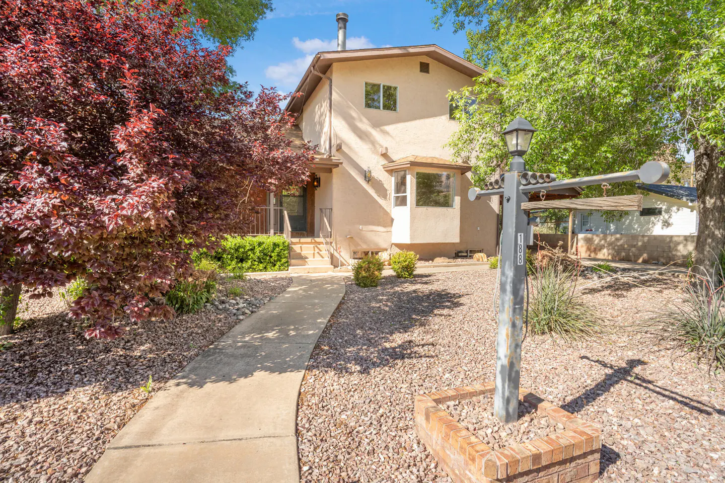Two-story beige house with a chimney, bay window, and a walkway leading to the front door. A red-leafed tree is on the left.