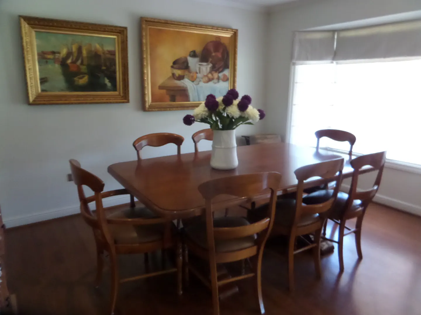 Dining room with a wood table, chairs, and hardwood floors. Two framed paintings hang on the wall. A vase of flowers sits on the table.
