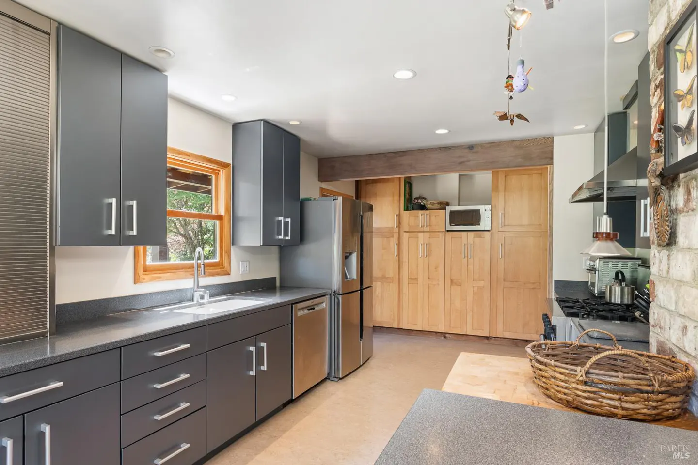 A modern kitchen with gray cabinets, stainless steel appliances, and light wood cabinets. A window provides natural light.
