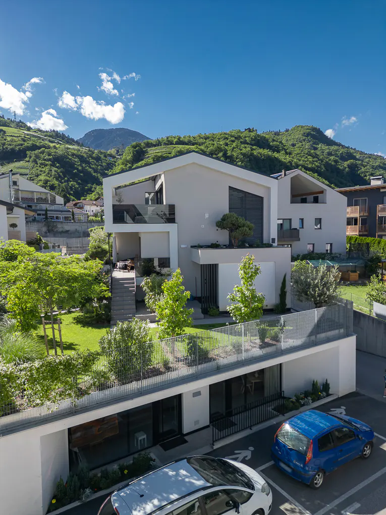 Modern white house with a green lawn, trees, and a mountain backdrop under a blue sky. Cars are parked in front.