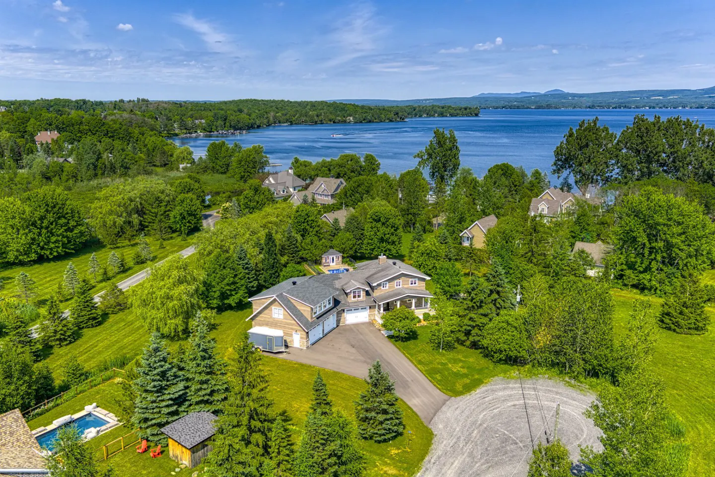 Aerial view of a large house with a driveway, pool, and lush green trees, situated near a scenic lake under a blue sky.