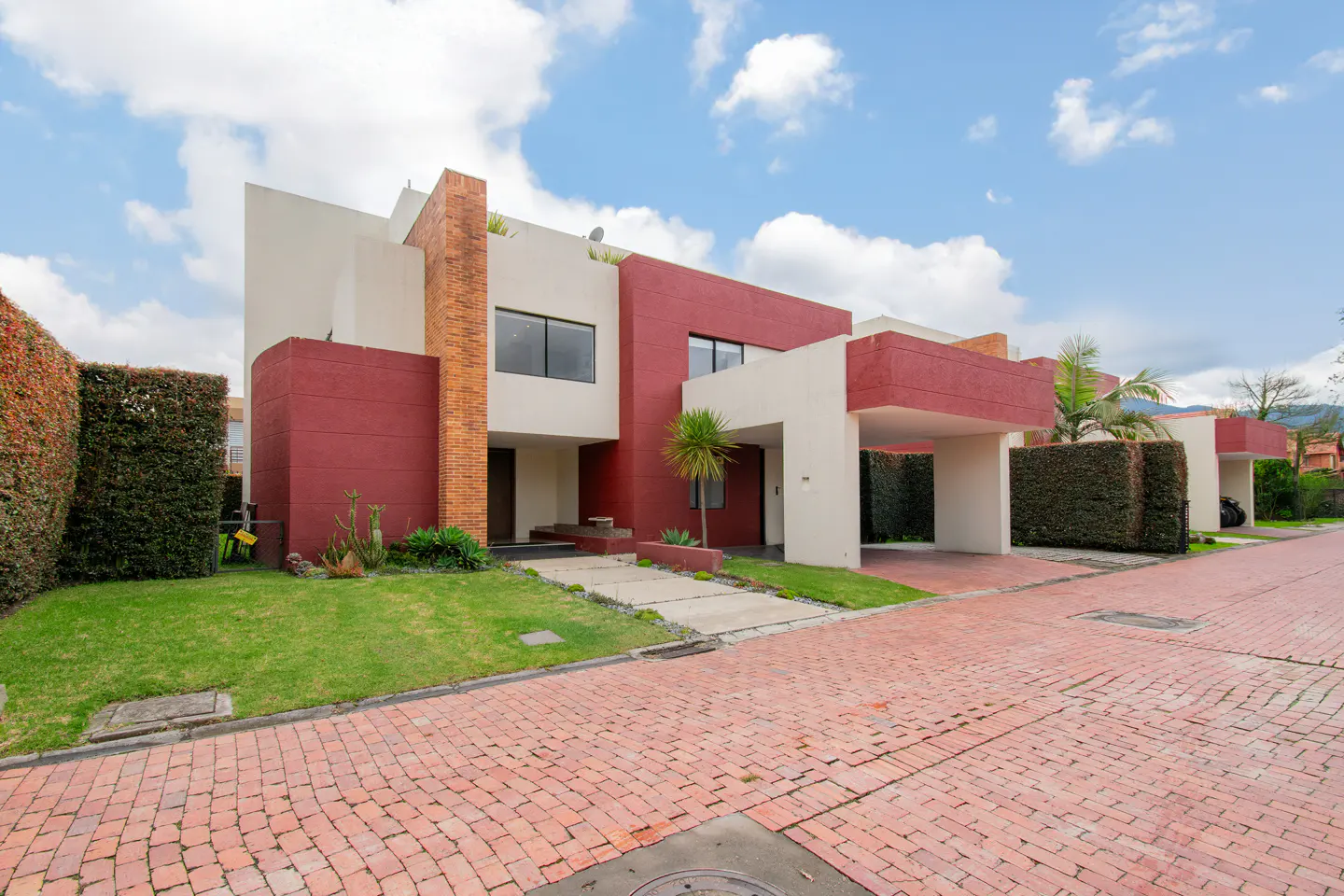 Modern two-story house with red and cream walls, a brick accent pillar, a carport, and a green lawn on a brick street.