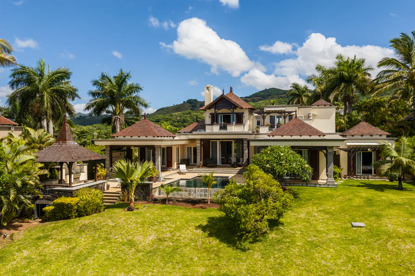 Luxury home with a pool and gazebo on a green lawn, surrounded by palm trees and lush vegetation under a blue sky.