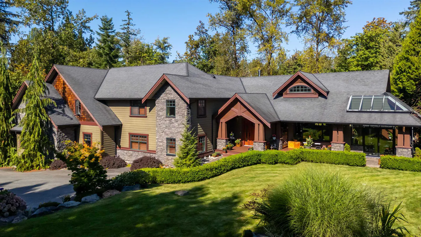 Large two-story house with gray roof, stone and olive green siding, surrounded by green lawn and trees under a blue sky.