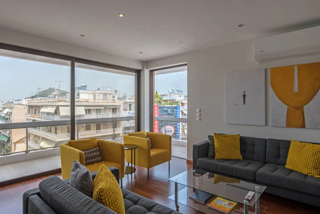 Bright living room with yellow armchairs, gray sofas, and a glass coffee table. Large windows offer a city view. Modern art hangs on the white walls.