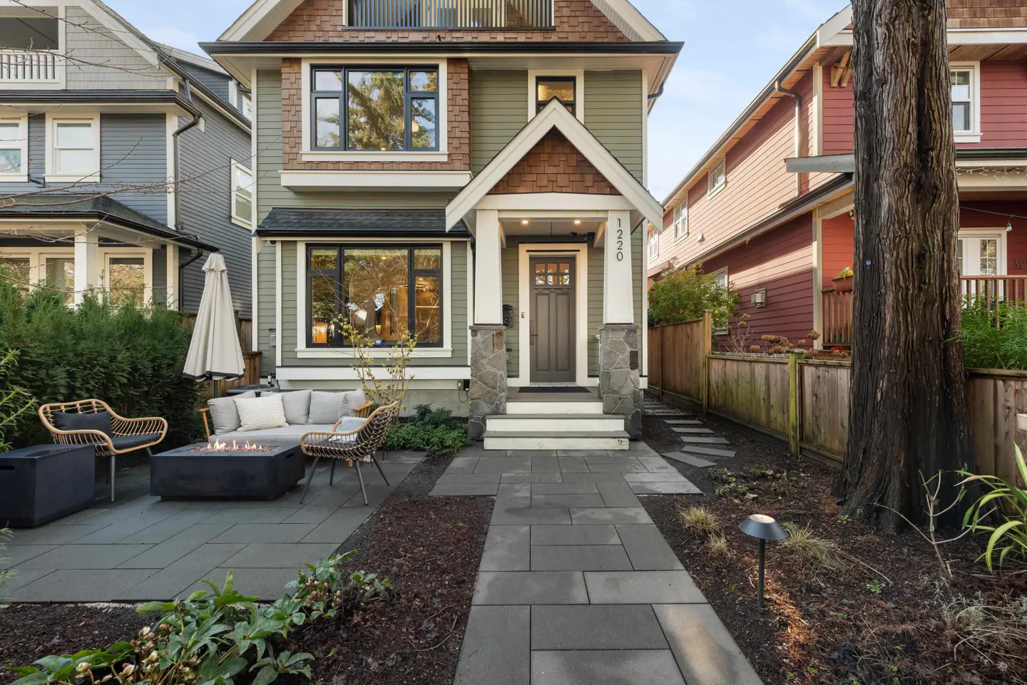 Two-story house with green siding and a brown shingle roof. A stone walkway leads to the front door. Outdoor seating area with a fire pit.