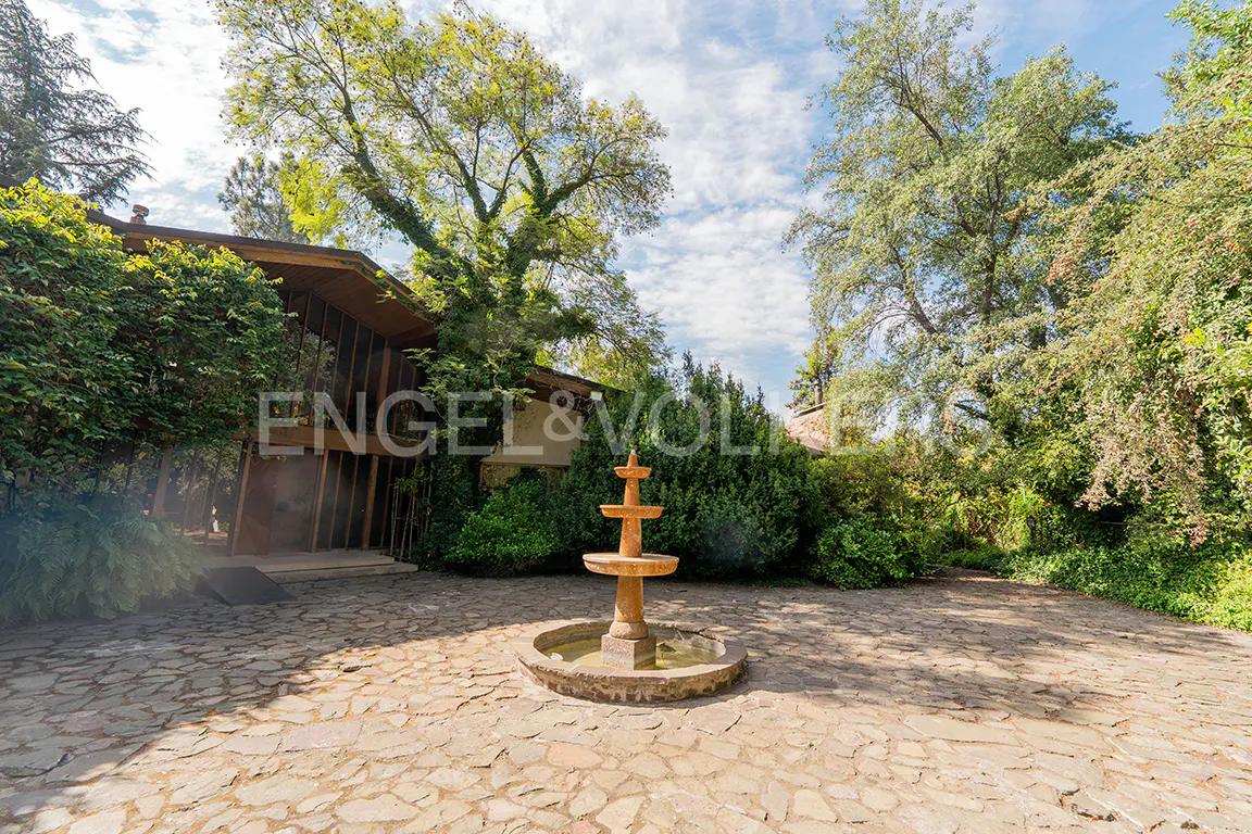 Exterior view of a home with a stone courtyard and a three-tiered fountain, surrounded by lush greenery and trees.