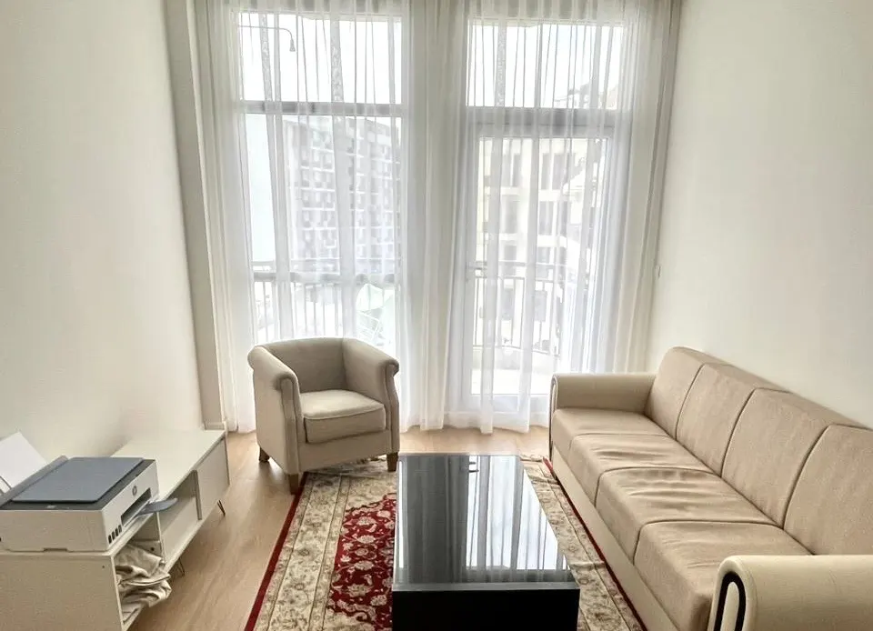 Living room with beige sofa and armchair, black coffee table, and red patterned rug. White walls and sheer curtains.