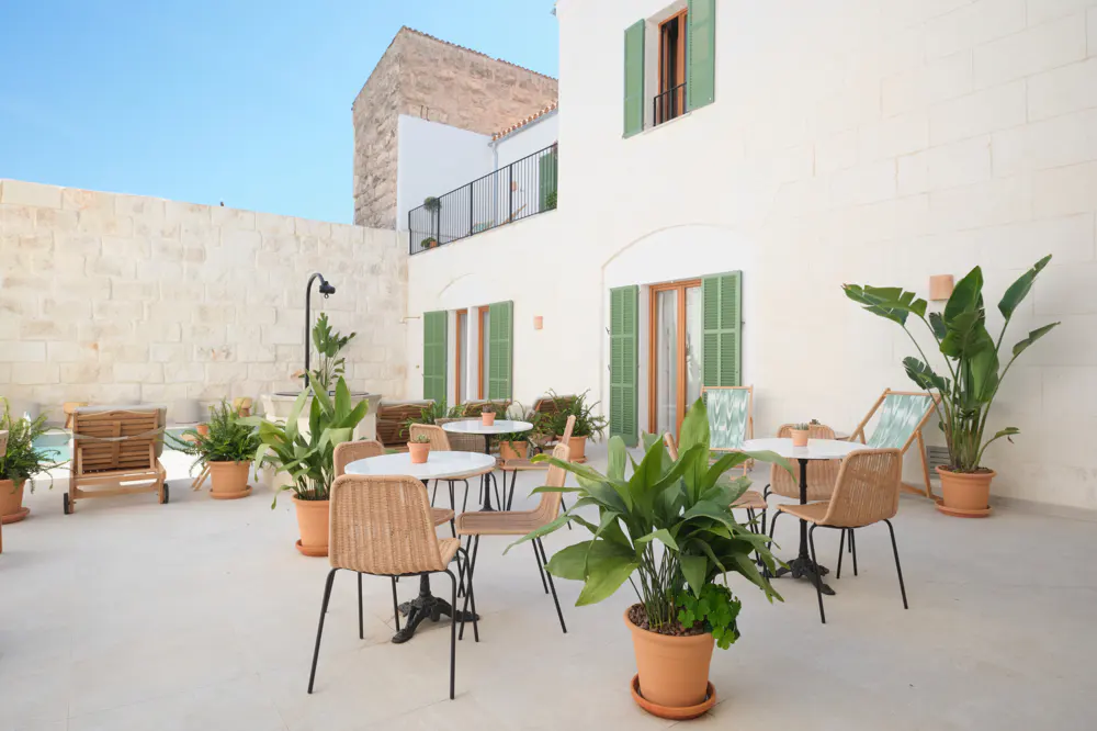 Outdoor patio with white walls, green shutters, and potted plants. Wicker chairs surround small tables. Lounge chairs are visible in the background.