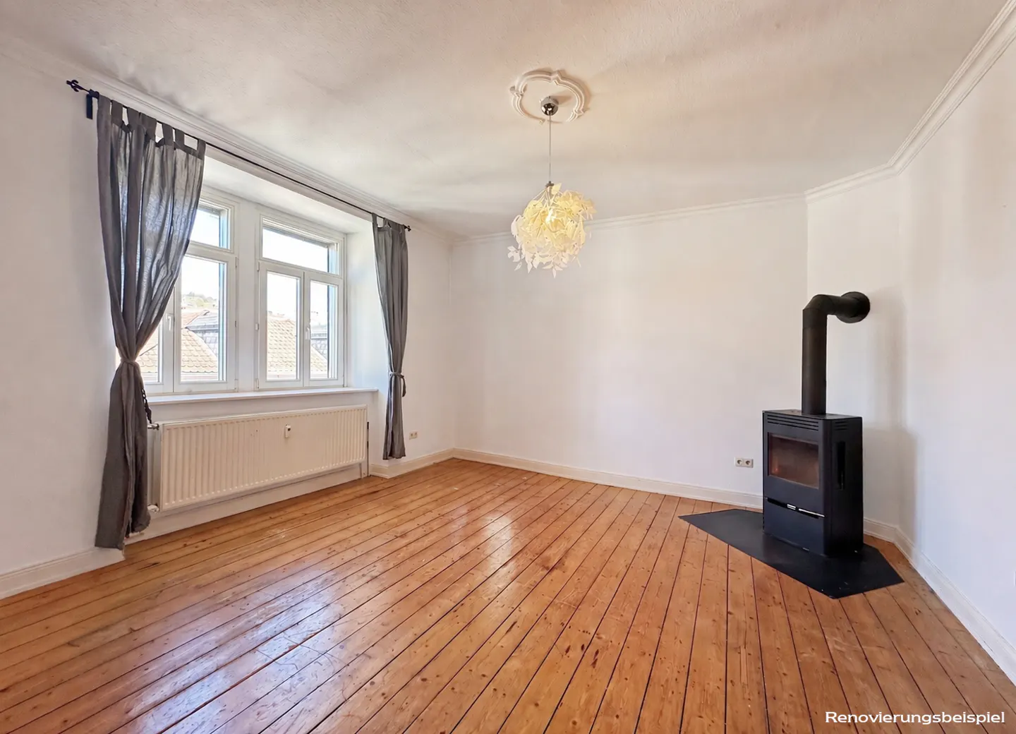 Bright room with wood floors, white walls, and a black wood-burning stove. Gray curtains frame a window with a radiator below. A decorative light fixture hangs from the ceiling.