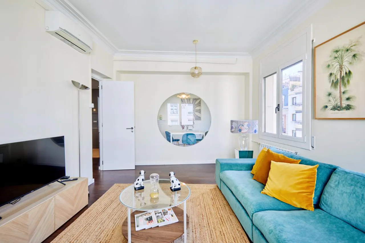 Bright living room with a teal sofa, yellow pillows, and a round window showing a dining area. A glass coffee table sits on a jute rug.