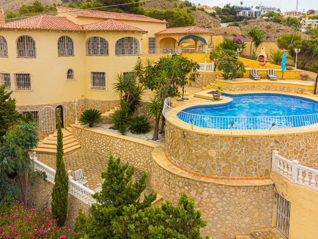 Yellow Mediterranean villa with a curved pool, stone retaining walls, lush landscaping, and a patio in a hilly setting.