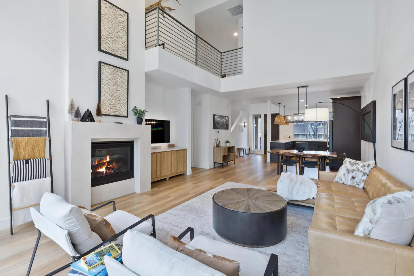 Bright, open-concept living room with a fireplace, tan leather sofa, and white chairs. A black metal staircase is visible in the background.