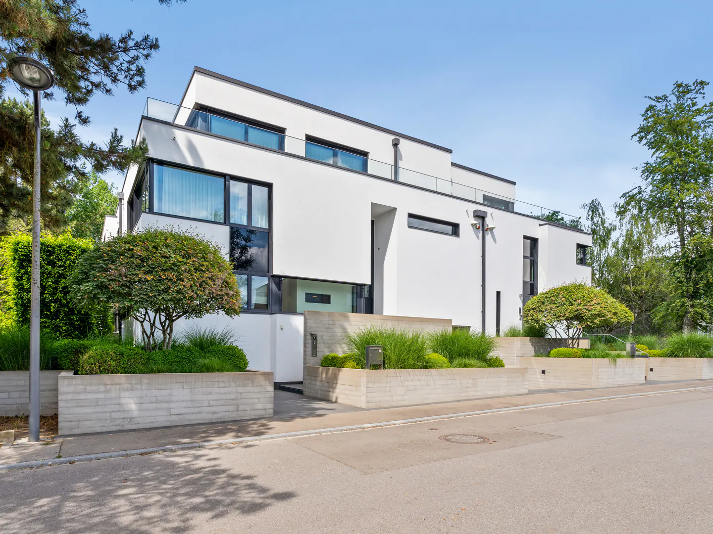 Modern white multi-story home with large windows, concrete planters, and manicured landscaping under a clear blue sky.