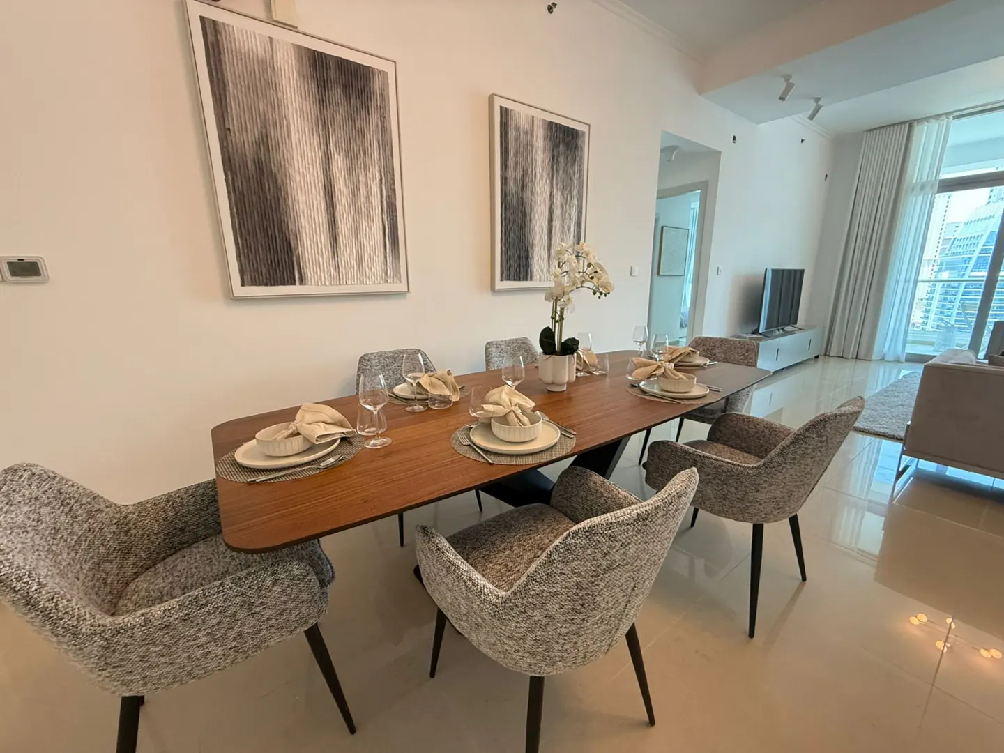 Dining room with a long wooden table set for a meal, surrounded by gray upholstered chairs. Two framed art pieces hang on the white wall.