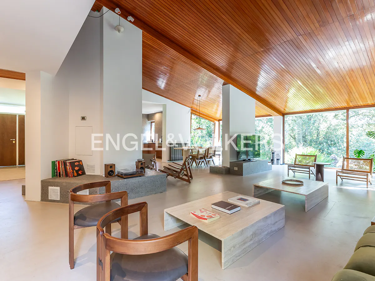 A bright living room with a wooden ceiling, stone coffee tables, and chairs. Large windows overlook a green outdoor space.
