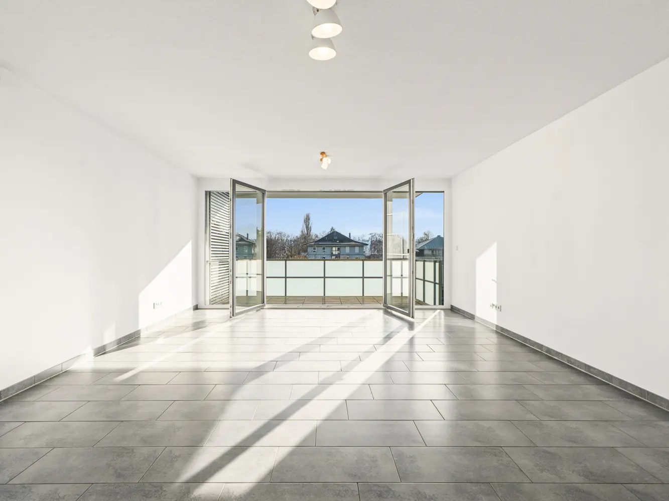 Bright, empty room with white walls and gray tile floor. Open glass doors lead to a balcony with a view of houses and trees.