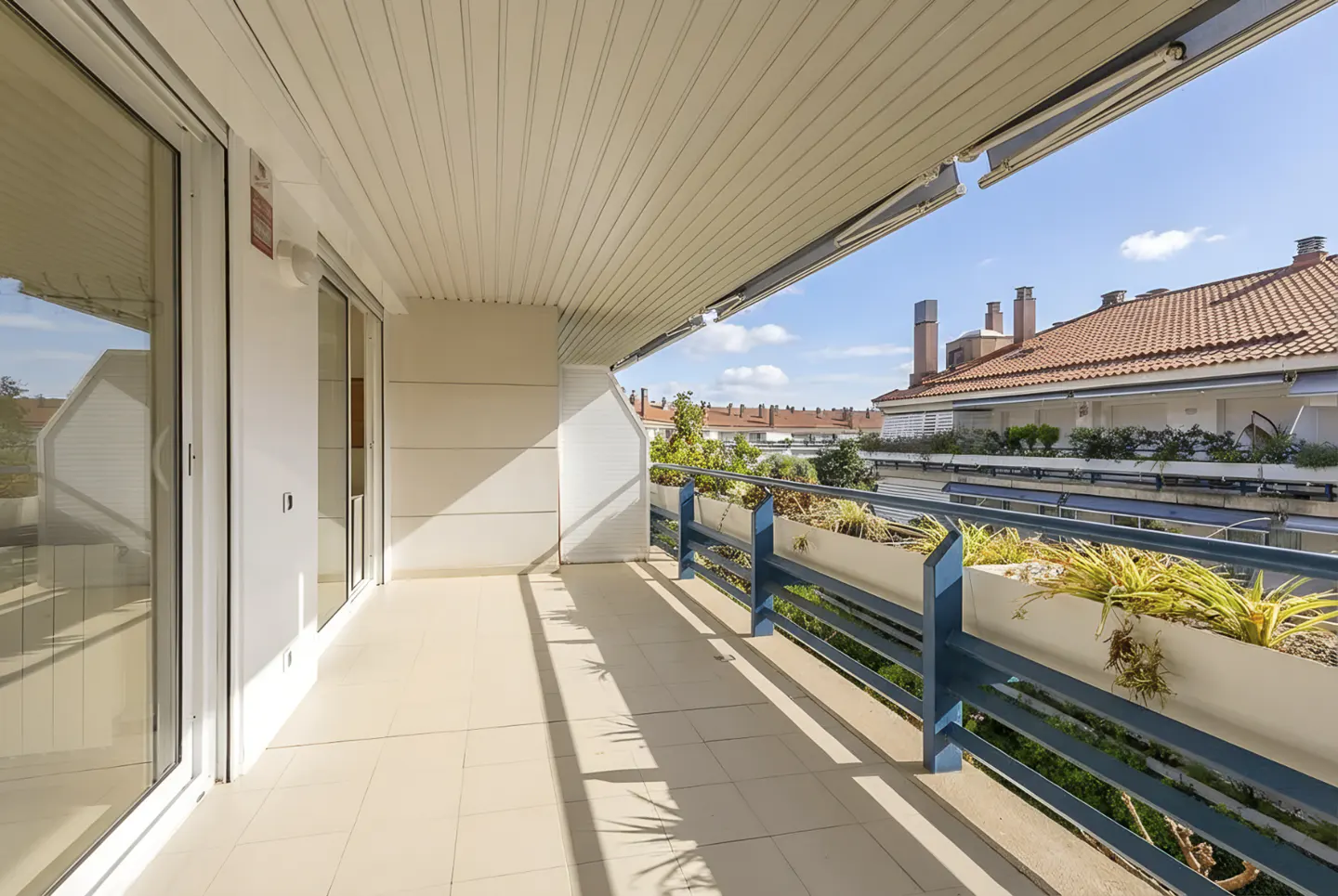 A bright, sunny balcony with blue railings and planters, overlooking rooftops with red tile roofs. Sliding glass doors lead inside.