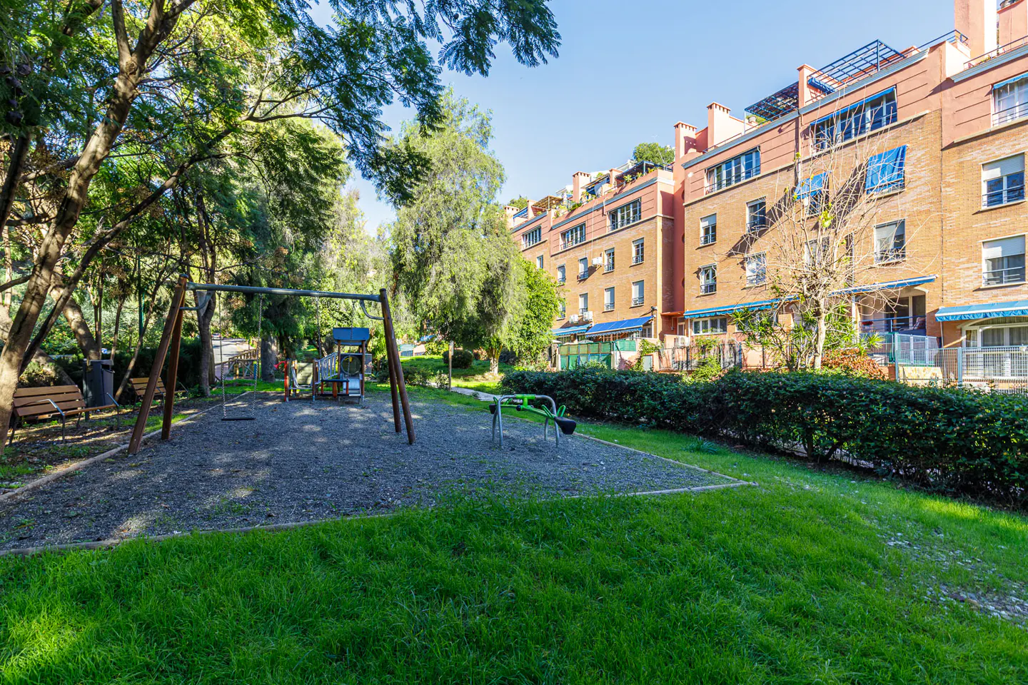 A playground with swings and a seesaw sits in front of brick apartment buildings with green trees.