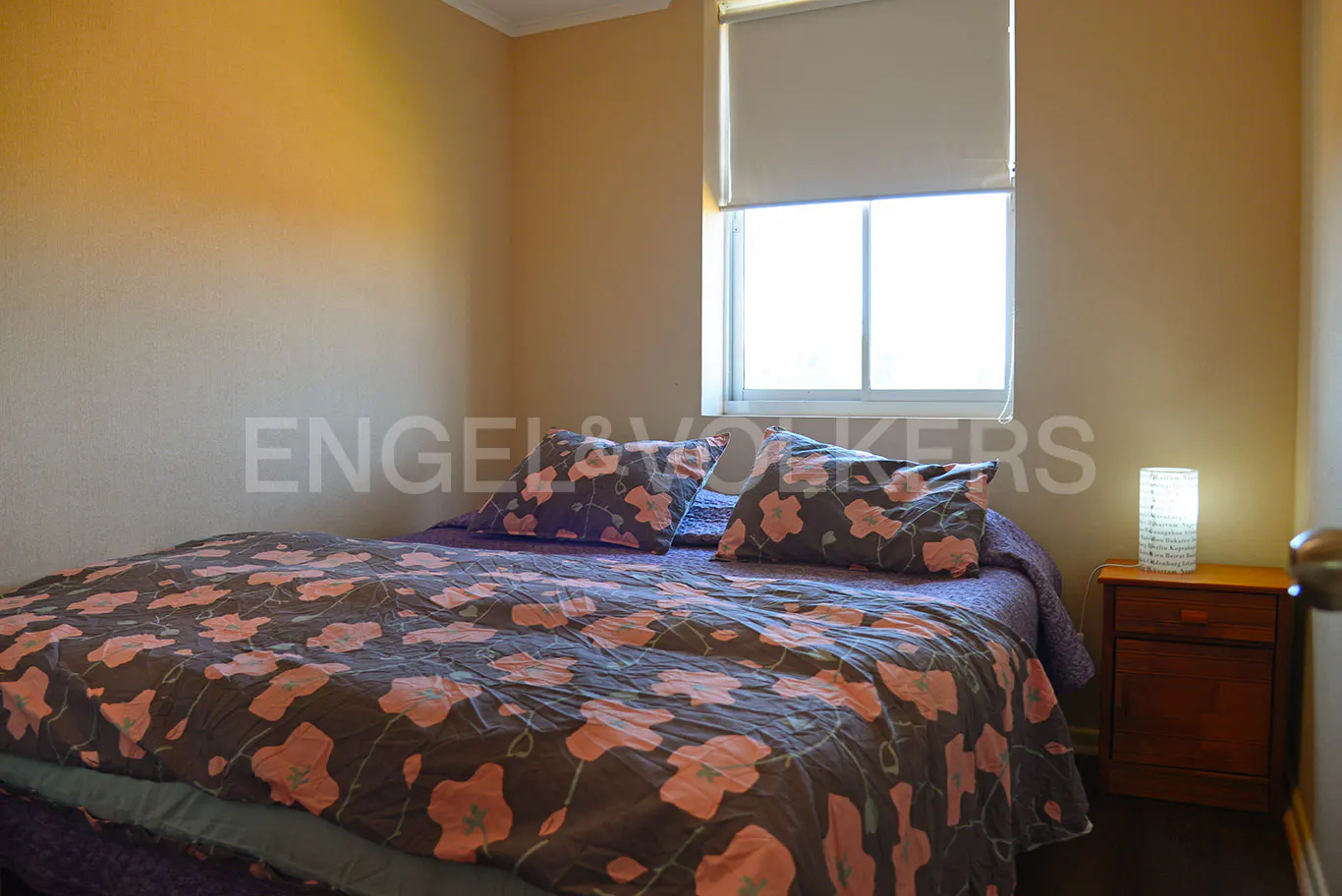 Bedroom with a bed covered in a brown and pink floral pattern, a window with a white shade, and a wooden nightstand with a lamp.