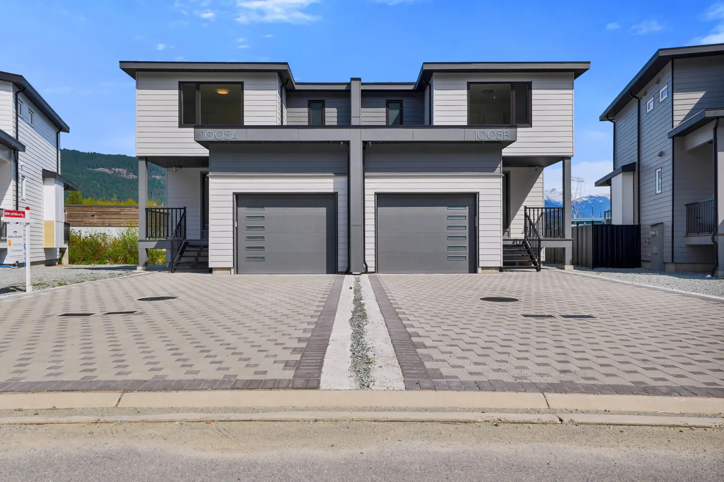 Two-story duplex with gray siding and garage doors, set against a blue sky and mountain backdrop. Paved driveway in front.