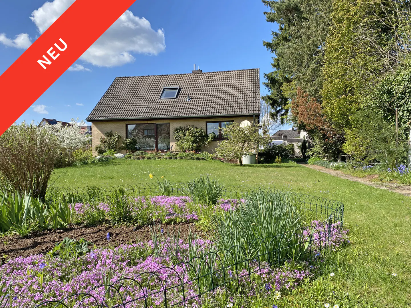 A single-story house with a brown roof and beige walls, surrounded by a green lawn and flowerbeds with purple flowers. A red banner with "NEU" is in the upper left corner.