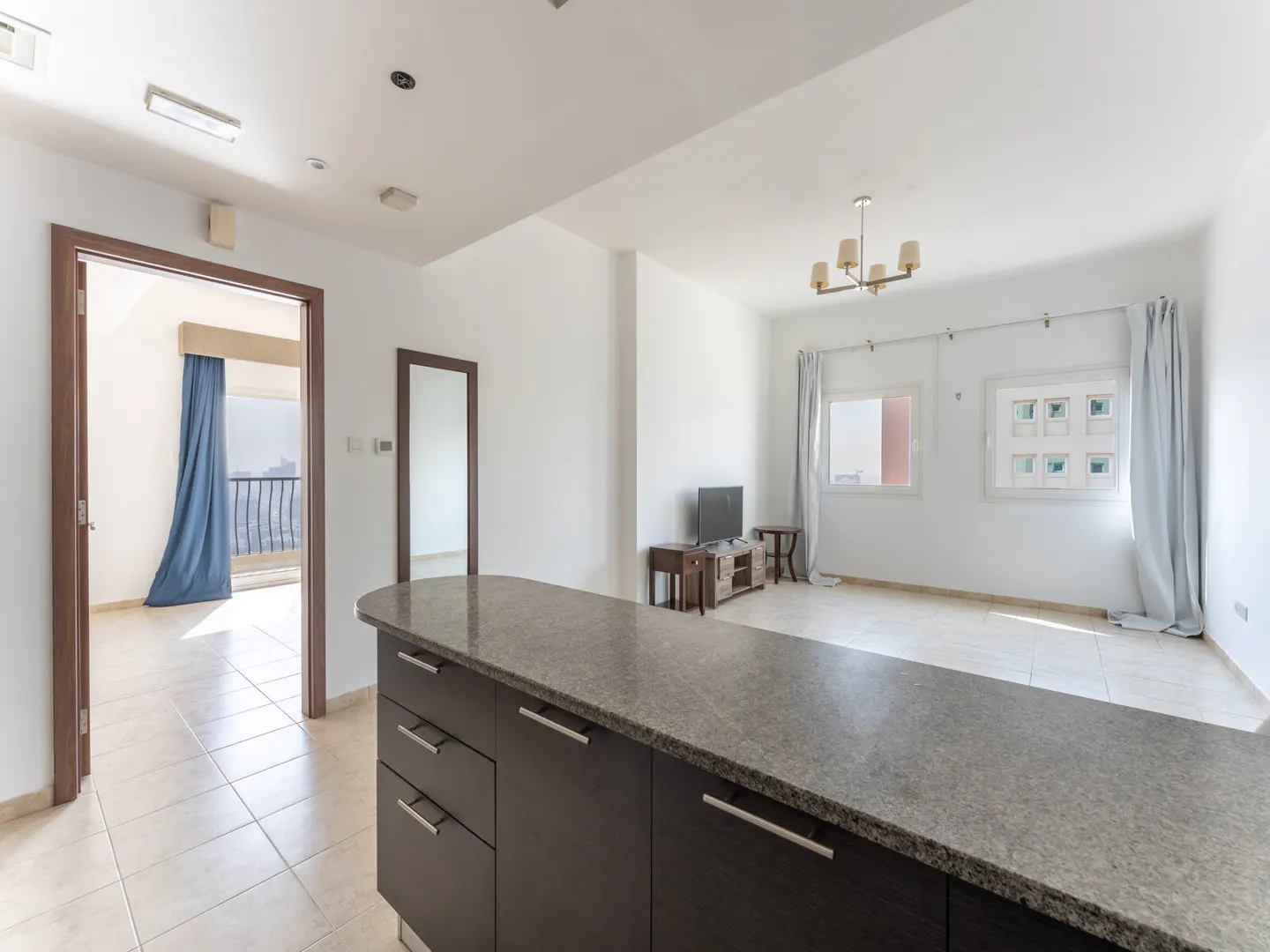 Open-concept apartment with a kitchen island, living area with TV, and balcony with blue curtains.