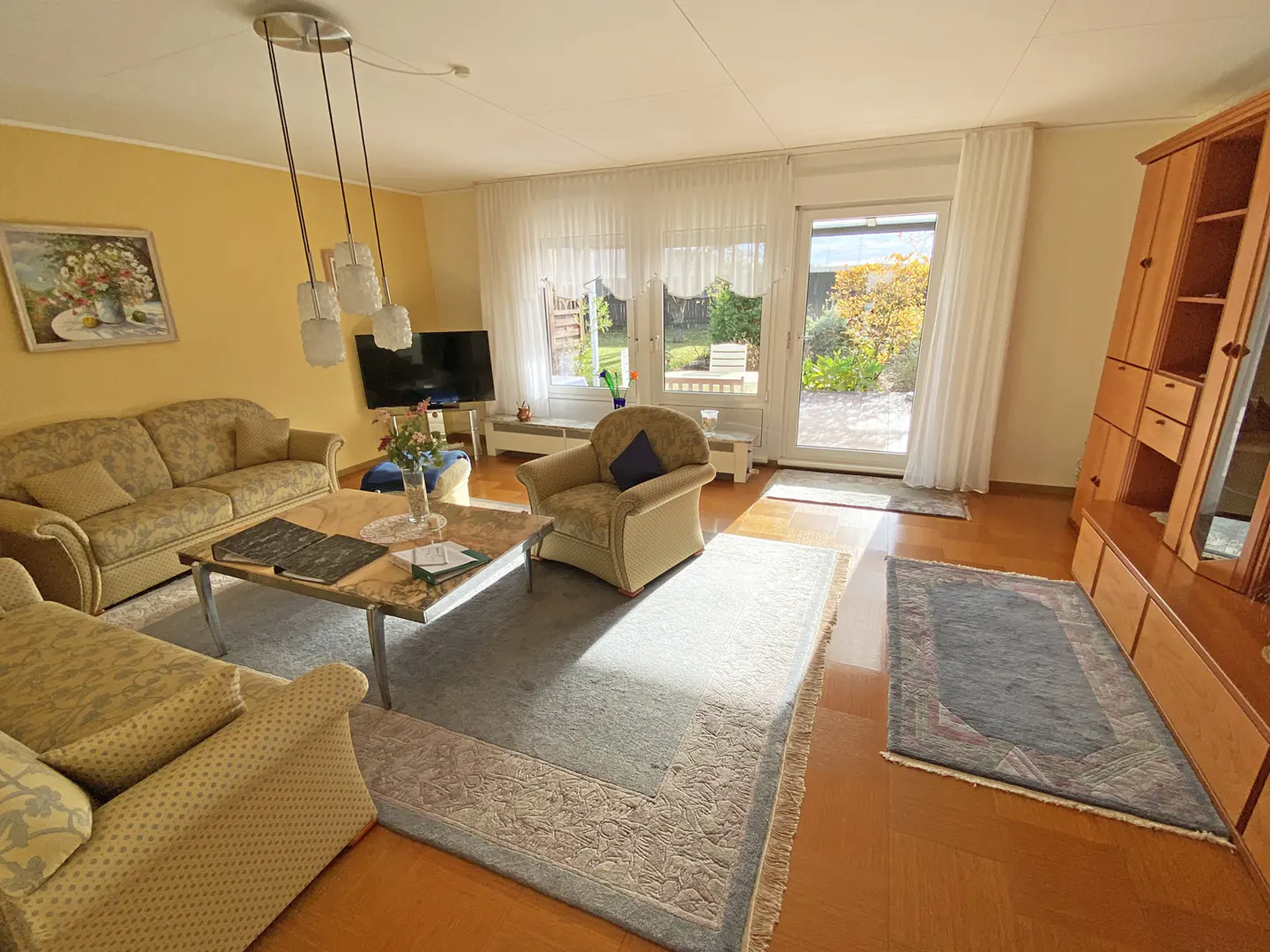 A sunny living room with patterned sofas, a marble-topped coffee table, and a view of the garden through sheer curtains.