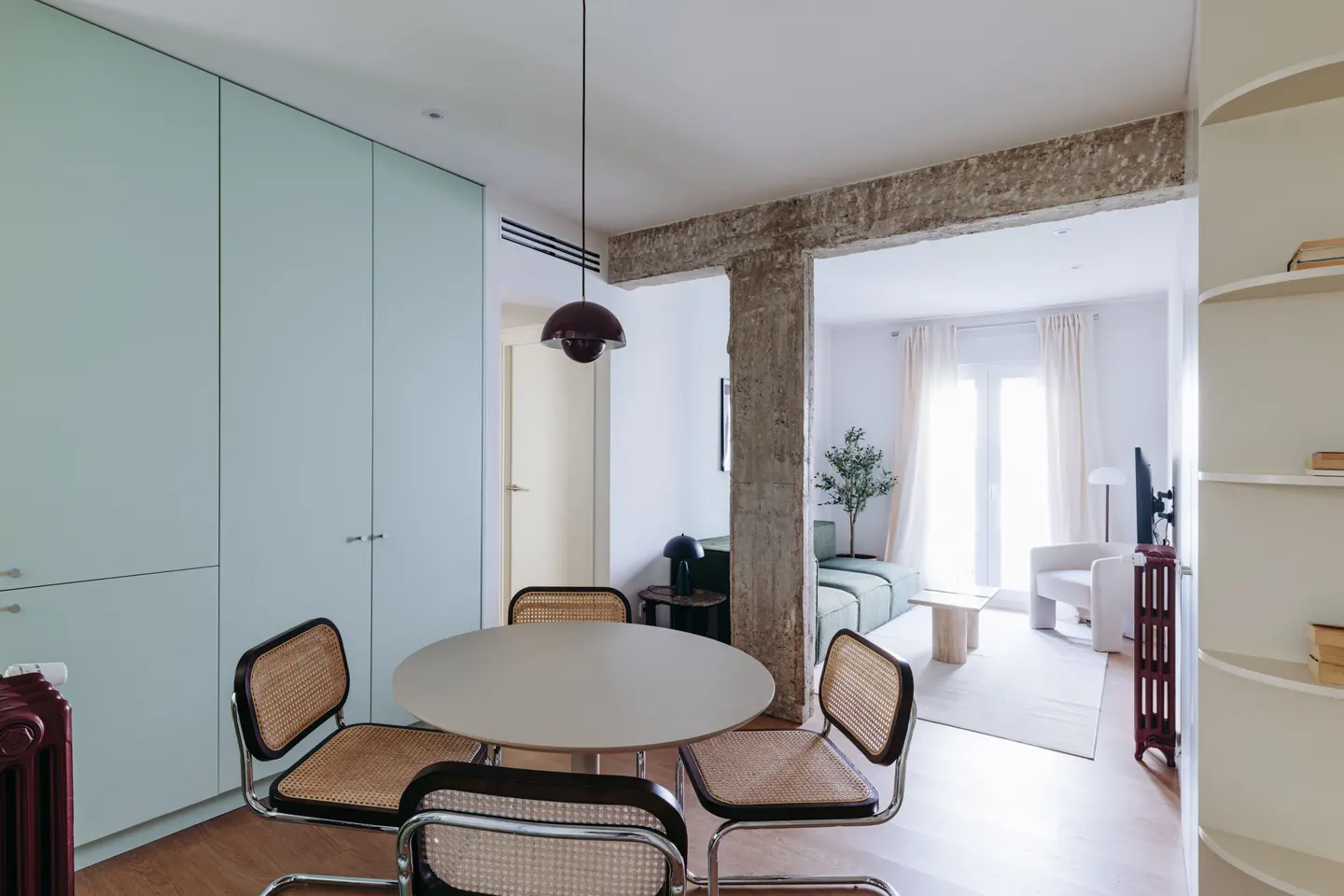 Bright apartment interior with a round table and four cane chairs. Living room visible through a concrete beam. Light blue cabinets.