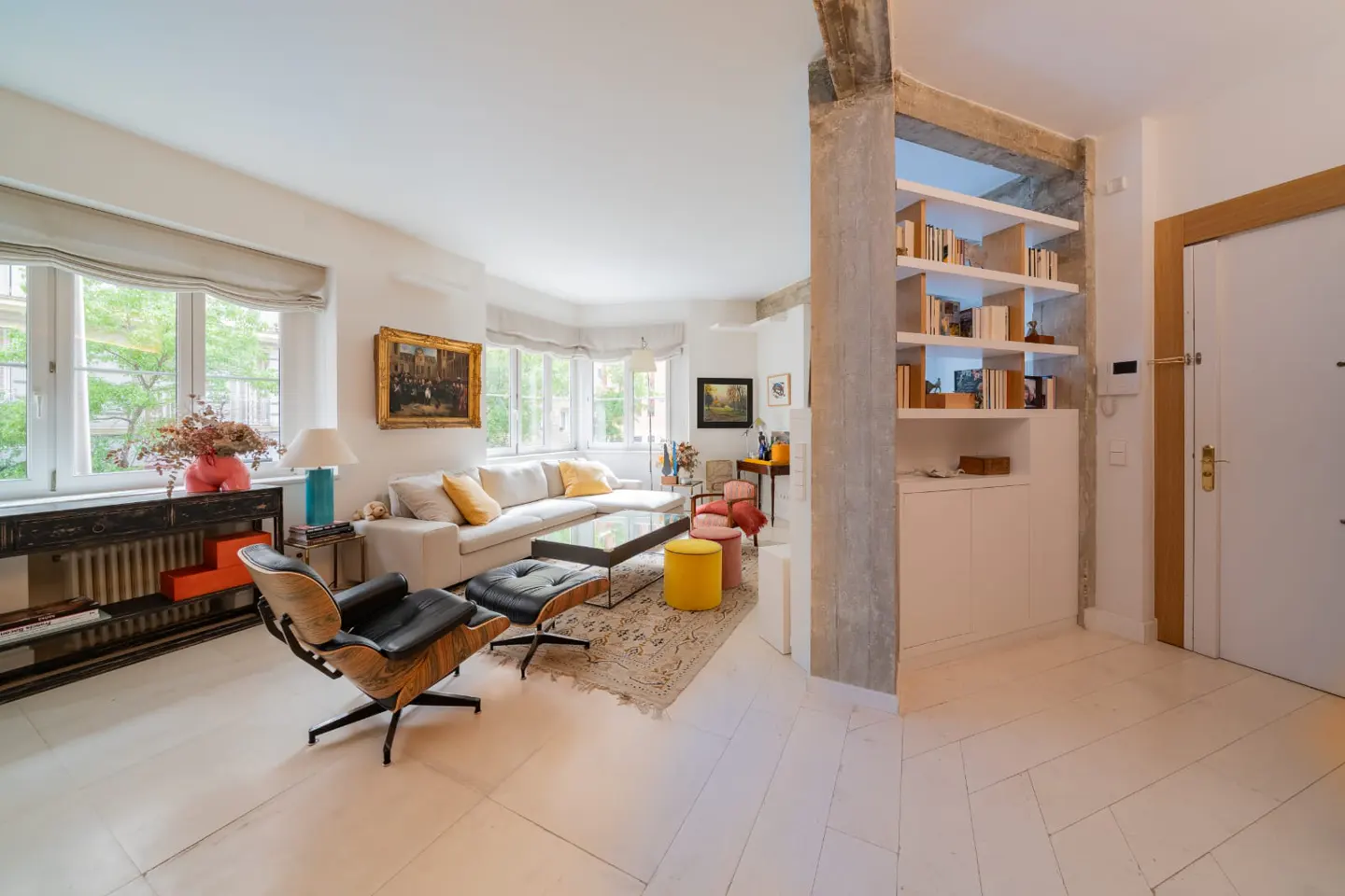 Bright living room with white wood floors, a cream sofa, and a black leather chair. A bookcase divides the room.