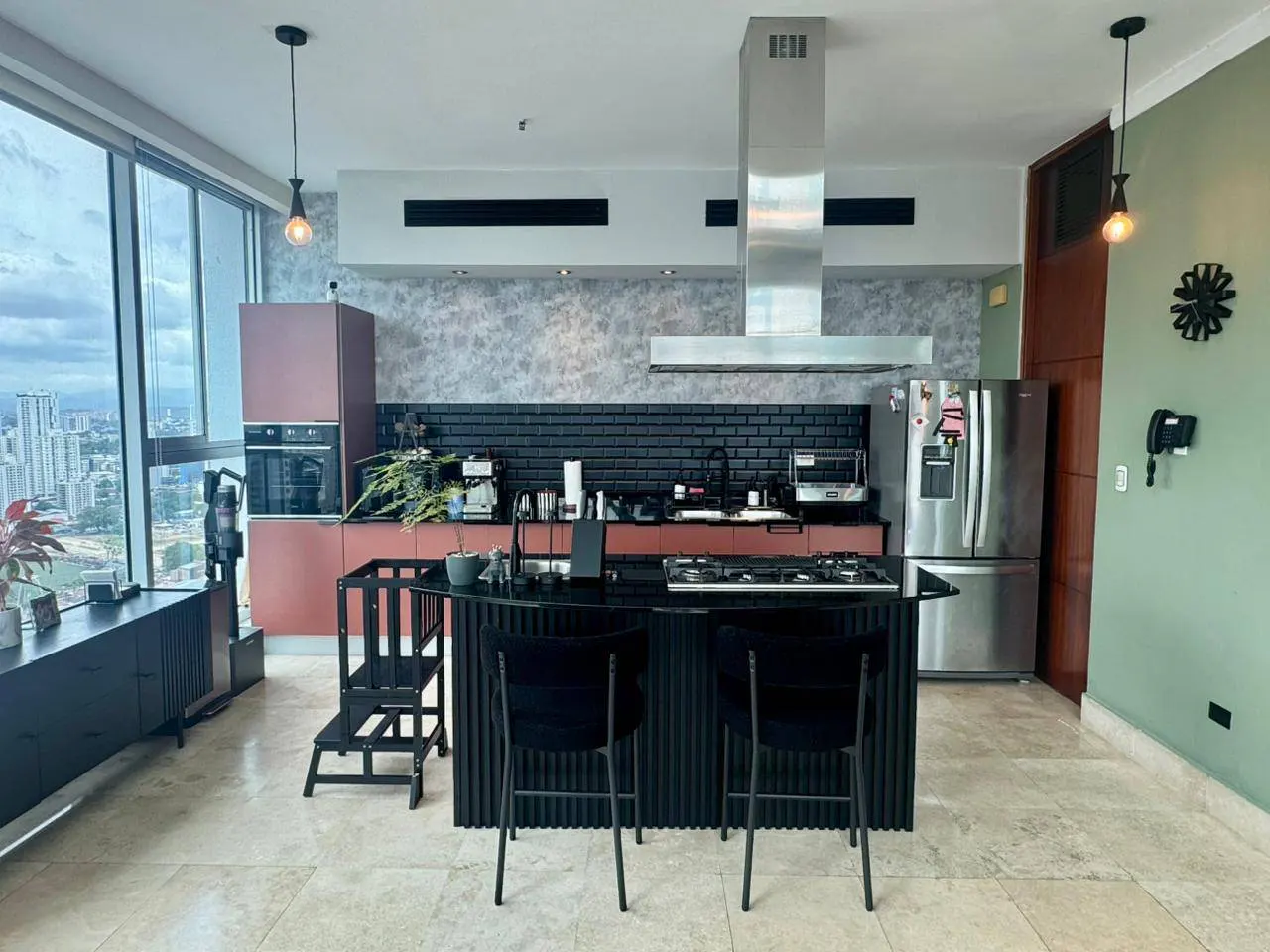 A modern kitchen with a black island, two black chairs, and stainless steel appliances. A large window shows a city view.