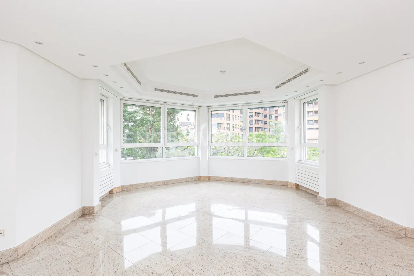 Bright, empty room with white walls, marble floors, and large windows. Outside, trees and buildings are visible.