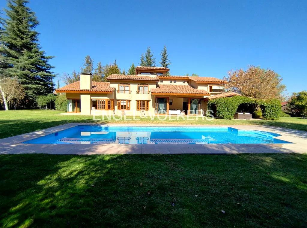 Exterior of a tan two-story house with a red tile roof, a blue pool, and green grass under a clear blue sky.