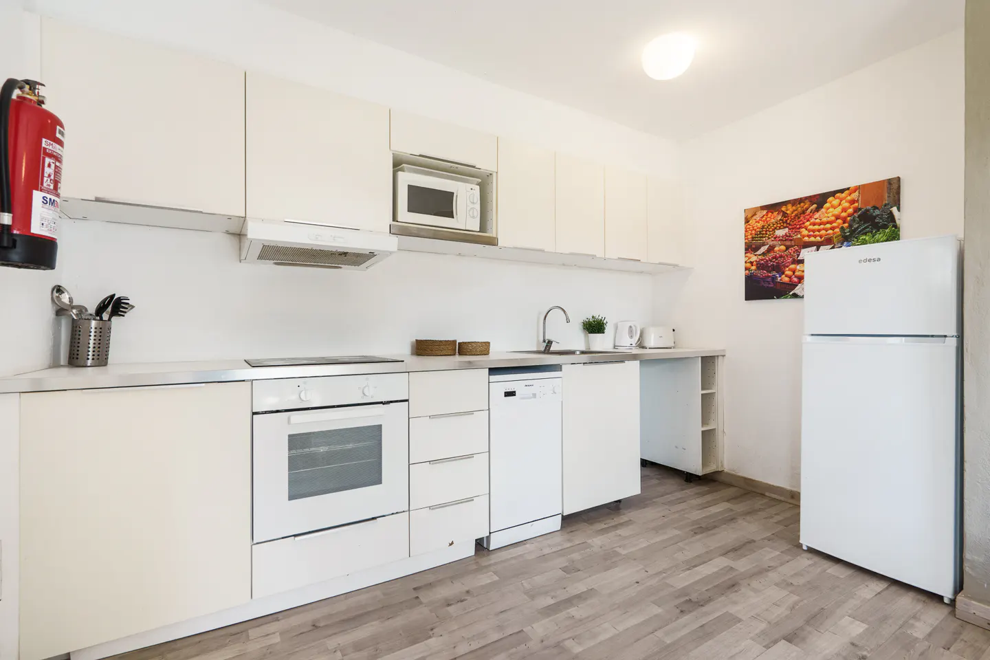 Bright, all-white kitchen with wood-look floors. Features oven, microwave, dishwasher, fridge, and fire extinguisher. Fruit art on the wall.