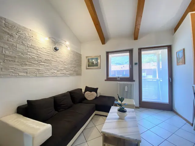 Living room with a dark brown sectional sofa, stone accent wall, and exposed wood beams. Windows and a door lead to a balcony.