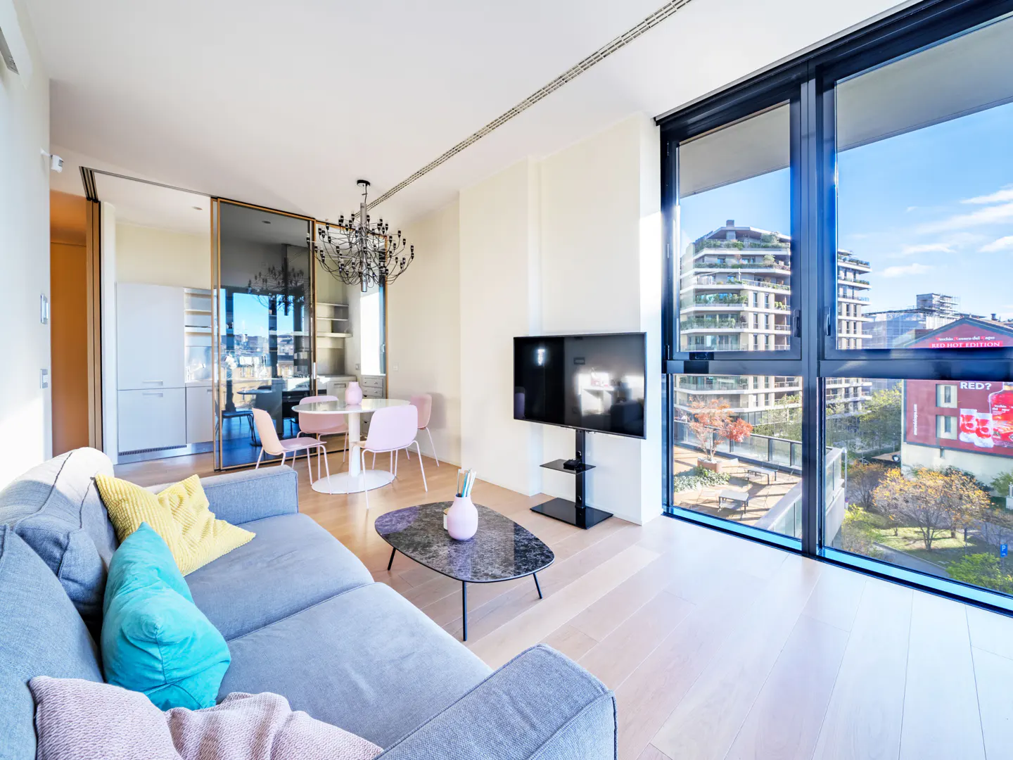 Bright apartment interior with a gray sofa, black coffee table, and TV. A dining area with pink chairs is visible through glass doors. Large windows offer a city view.