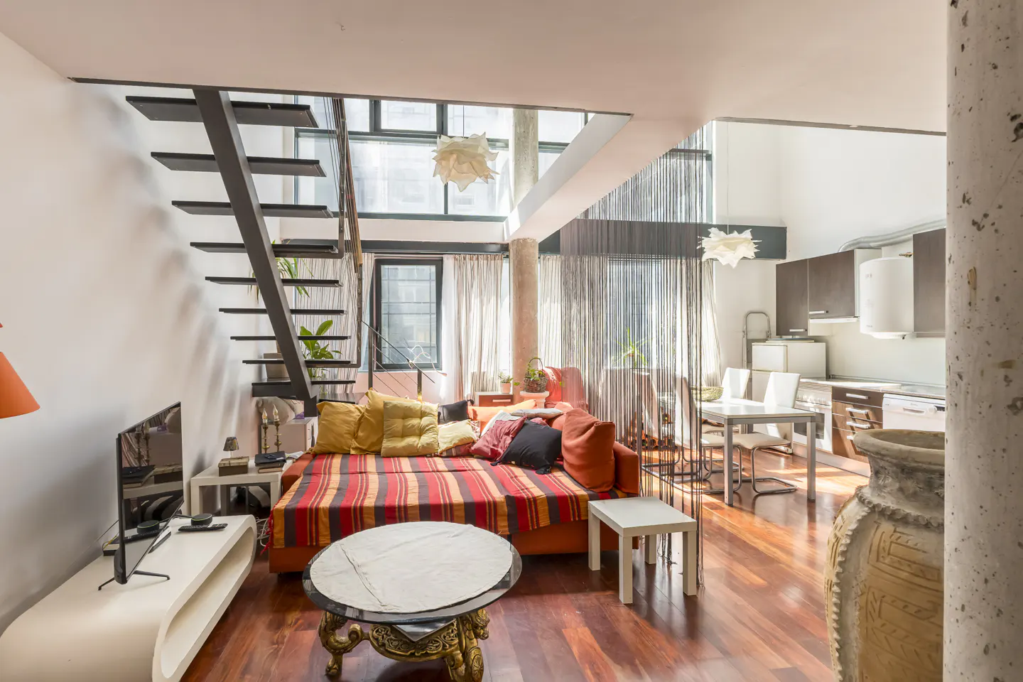 Bright, open-concept living space with hardwood floors, orange sofa with colorful striped blanket, black staircase, and kitchen in the background.