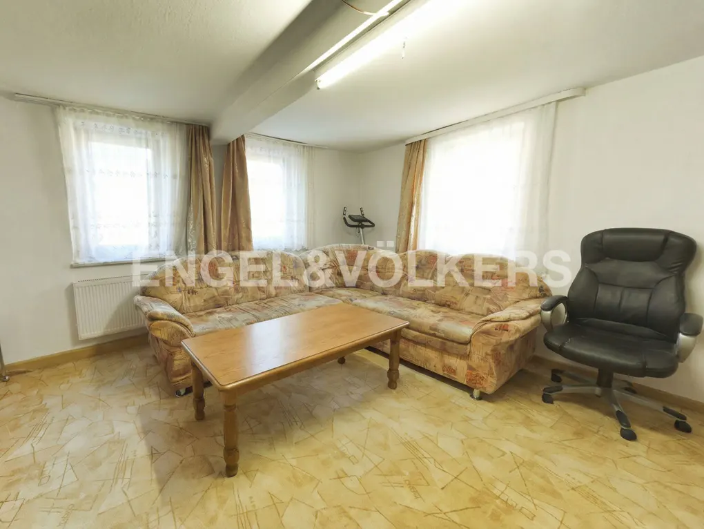 Living room with a patterned beige sectional sofa, wooden coffee table, black office chair, and windows with curtains.