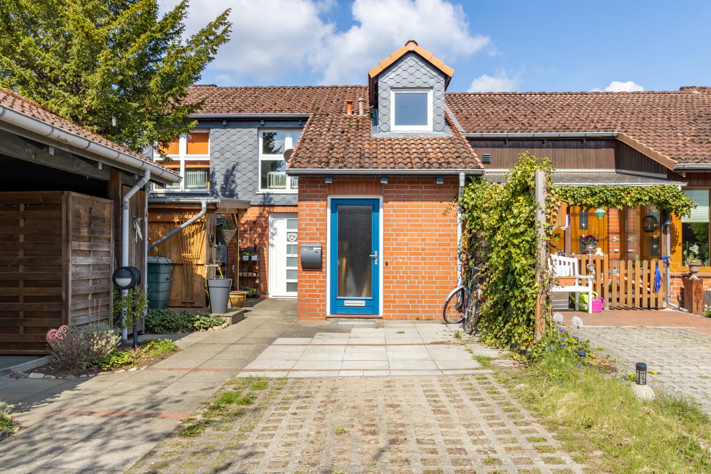 Exterior view of a row house with brick facade, blue door, and a driveway made of concrete blocks. A bicycle is parked on the side.