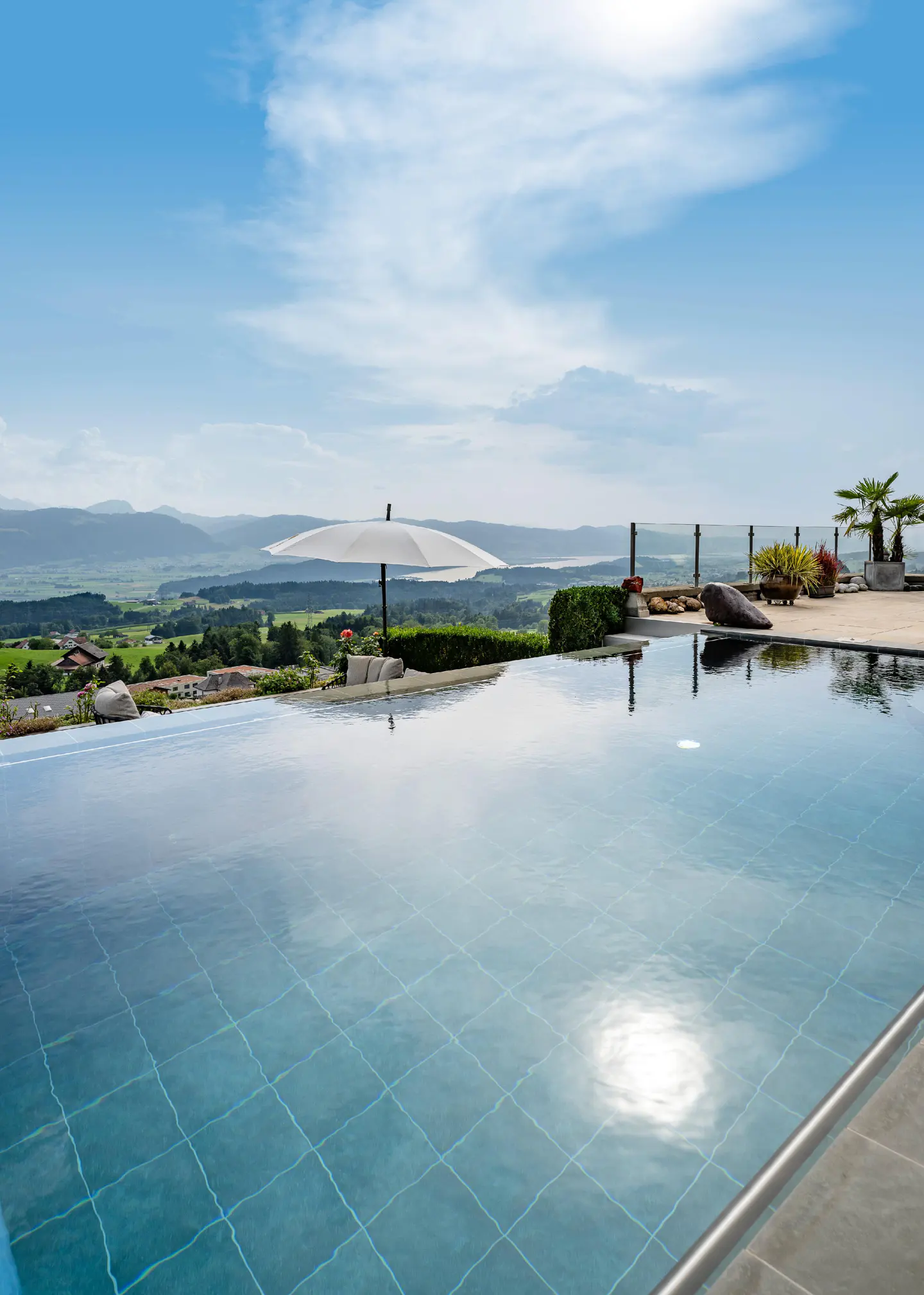 Infinity pool with blue tiles overlooks a green valley and mountains under a blue sky with white clouds.