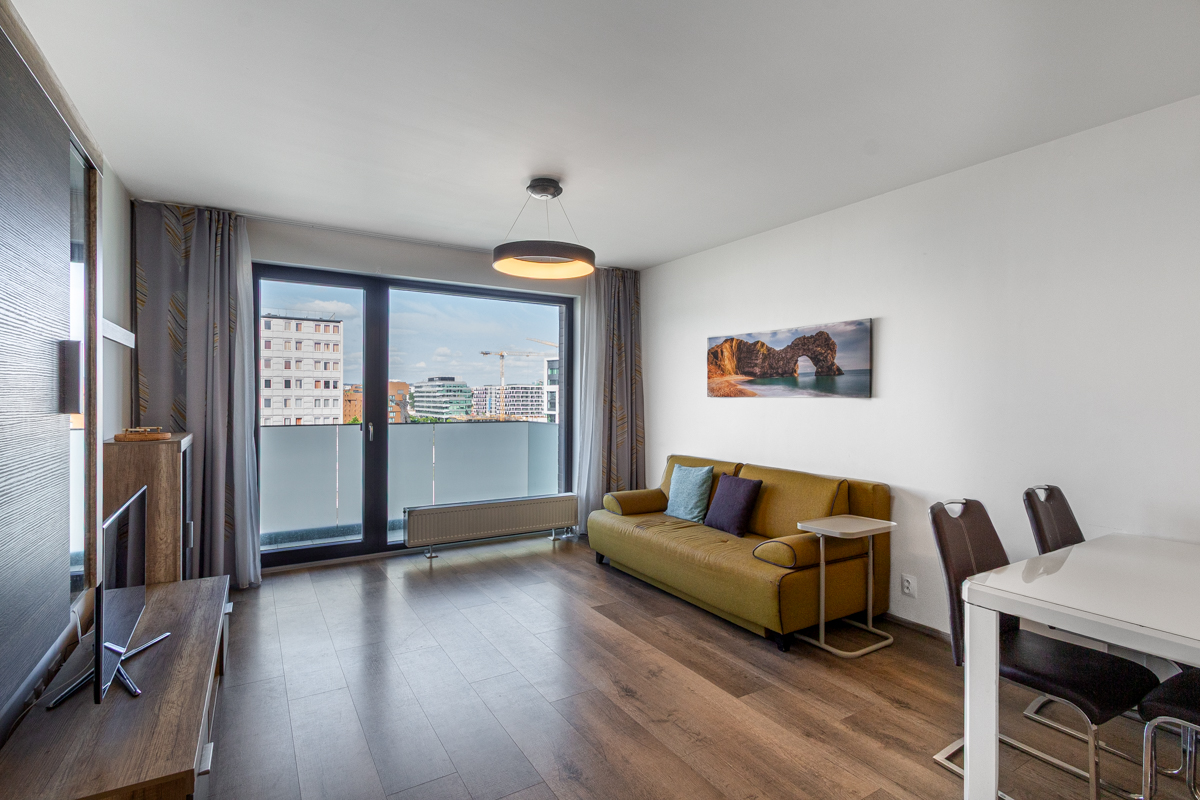 Modern living room with wood floors, mustard sofa, dining area, and a large balcony door overlooking a city skyline.