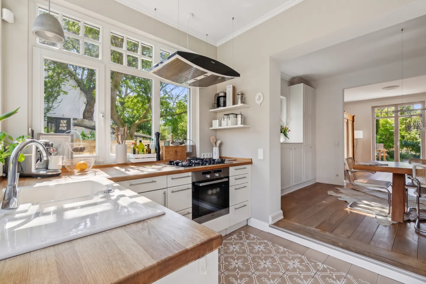 Bright kitchen with wood countertops, white cabinets, and a large window overlooking green trees. A dining room is visible in the background.