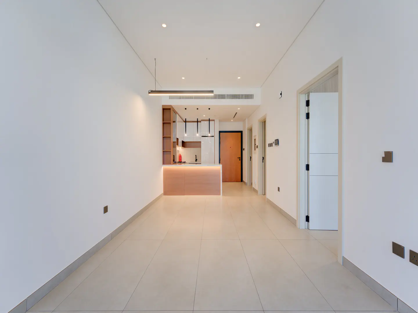 A bright, empty apartment with white walls, beige tile floors, and a modern kitchen with wood cabinets.