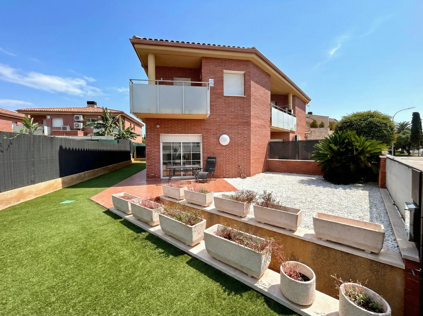 Two-story brick house with balconies, a patio with a table and chairs, and a landscaped yard with planters and white gravel.