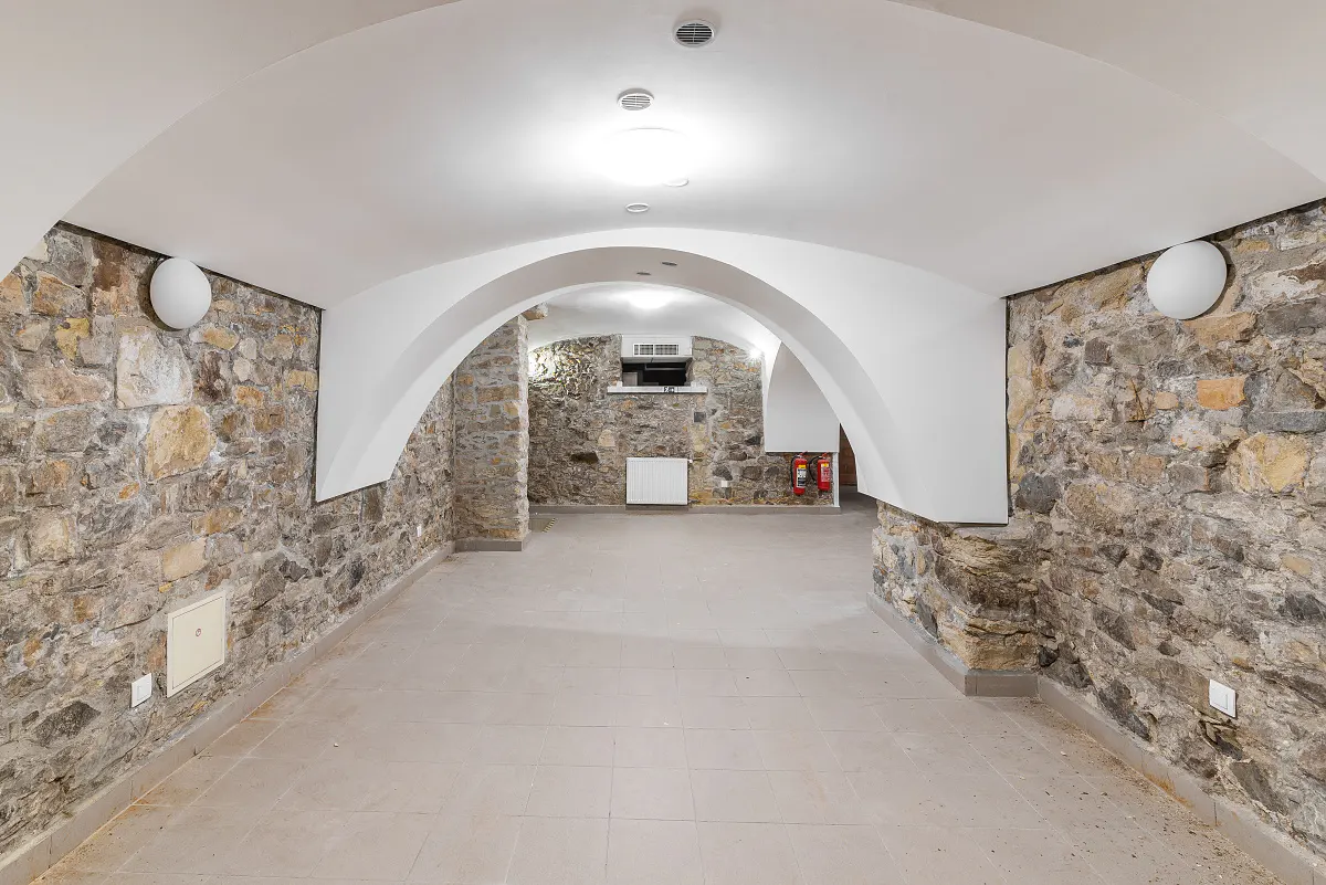 A basement room with stone walls, arched ceilings, and tile floors. White paint accents the arches and ceiling. Two red fire extinguishers are visible.