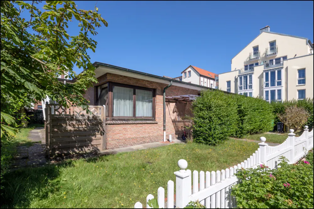 Brick house with a white picket fence and green lawn on a sunny day. Bushes and trees surround the house.