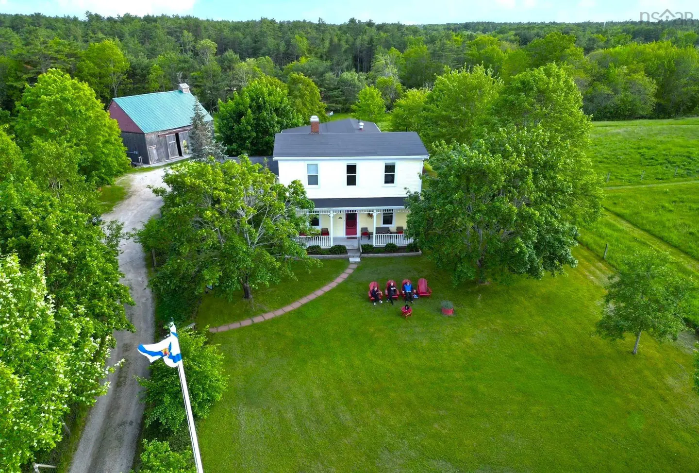 Aerial view of a yellow two-story house with a red door, surrounded by green trees and lawn. People sit in red chairs. A barn and Nova Scotia flag are visible.