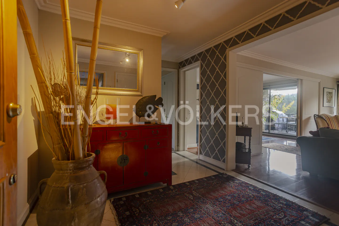 Entryway with a red cabinet, a vase with bamboo, and a patterned rug. A mirror hangs above the cabinet. The Engel & Völkers logo is visible.