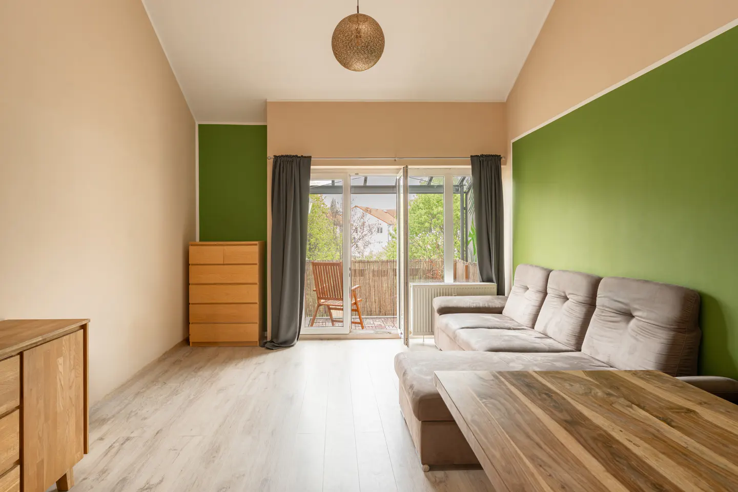 Living room with beige walls, green accent walls, light wood floors, and a brown sectional sofa. Balcony with chairs visible.